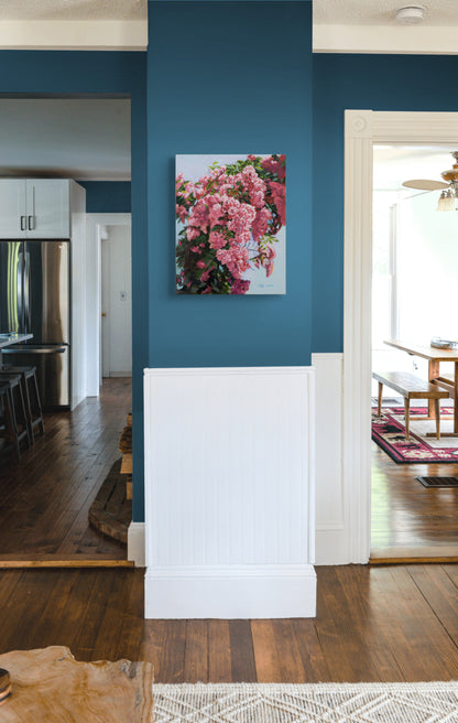 A vertical canvas wall art print depicting a dense cluster of pink and red flowers, displayed on a dark blue wall above white wainscoting in a home interior.